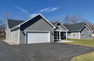 A modern, single-story gray house with a two-car garage, black shingled roof, and white trim, set on a paved driveway with a grassy lawn and leafless trees in the background under a clear blue sky.
