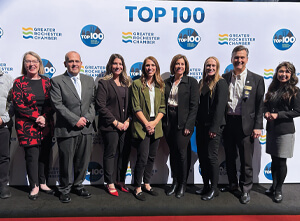 A group of nine people standing side by side, smiling, in front of a Greater Rochester Chamber Top 100 step-and-repeat banner at an event.