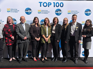 A group of nine people standing in front of a step-and-repeat banner displaying TOP 100 and Greater Rochester Chamber logos, posing and smiling for a photo at an event.