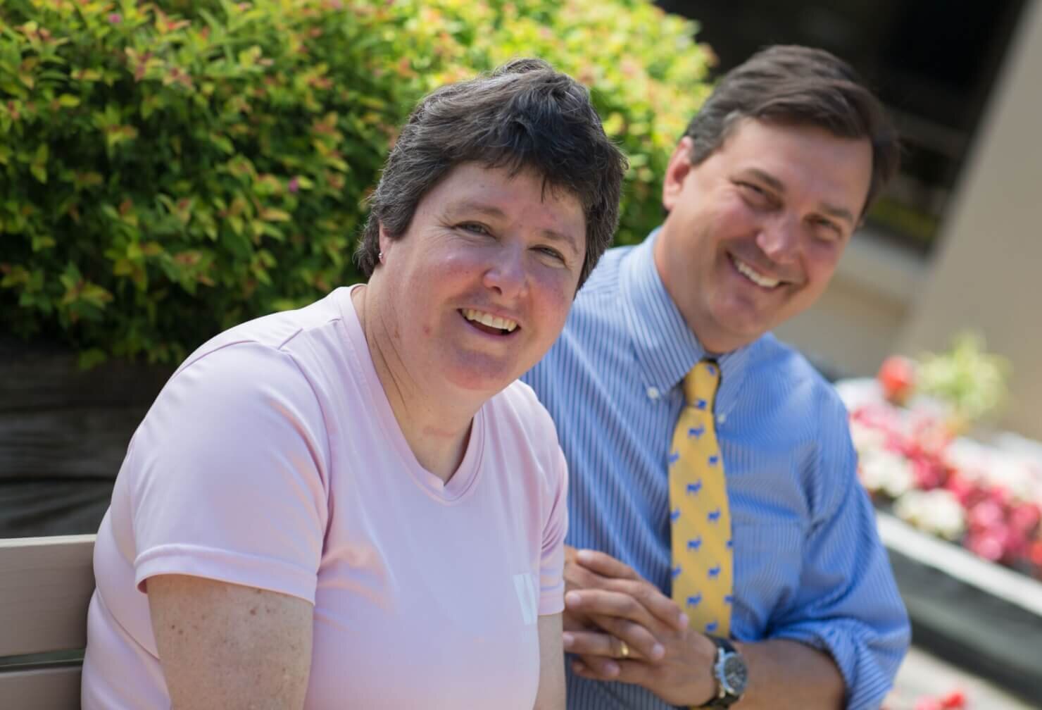 Two adults smiling while sitting outdoors on a bench, with flowers and greenery in the background. The person in front wears a light pink shirt, and the person behind wears a striped shirt with a yellow tie.