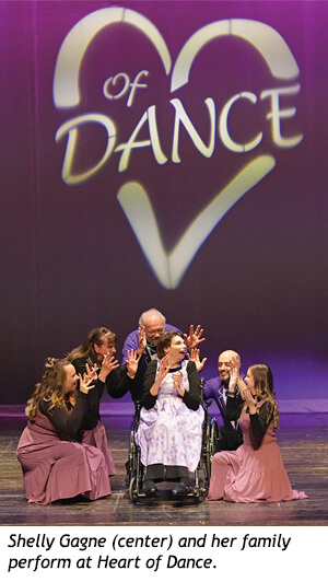 Shelly Gagne, seated in a wheelchair, and her family perform on stage in front of a backdrop featuring a large heart logo with the text Of DANCE.