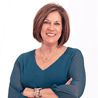 A smiling woman with shoulder-length hair wearing a teal long-sleeve v-neck top with her arms crossed, for the Fall 2026 Spirit Newsletter.