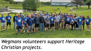 A large group of volunteers in matching blue shirts standing in a grassy park, with text that reads, Wegmans volunteers support Heritage Christian projects.